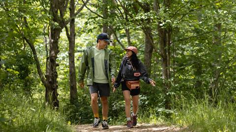 Two people hiking on a forest trail, smiling and conversing, dressed in casual hiking attire with backpacks.
