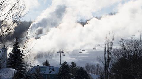 Snow-covered ski slope with active snow machines creating large clouds of artificial snow, ski lifts visible in the background under a clear sky.