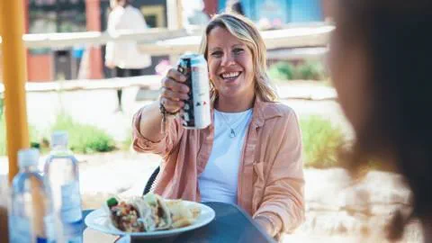 A woman drinking a can of beer at an outdoor table.