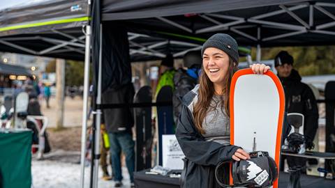 A joyful woman holding a snowboard at an outdoor equipment stall in a snowy environment.