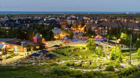 Aerial view of a resort village at dusk with illuminated buildings, a mini-golf course in the foreground, and a forest backdrop.