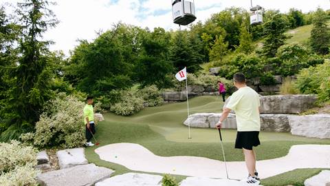 Two people playing mini golf on a course with cable cars overhead and rocky, green landscape around.