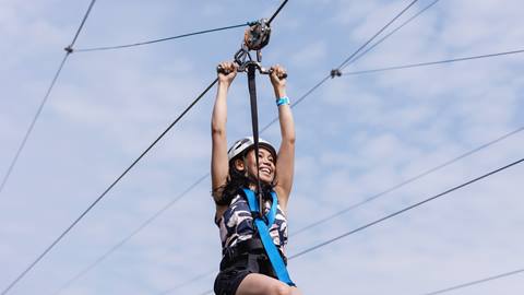 A woman wearing a helmet and harness joyfully zip lining against a clear blue sky.