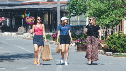 Three women walking on a sunny street, holding shopping bags and coffee cups, dressed in casual summer attire.