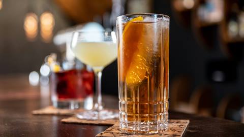 A tall glass of iced tea with a lemon twist on a bar, accompanied by other cocktail drinks in the background.