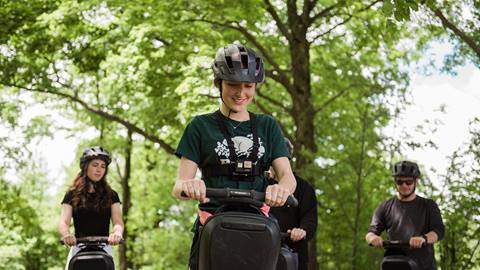 A young woman in a helmet smiles while riding a segway in a lush park, with two other people in helmets riding behind her.