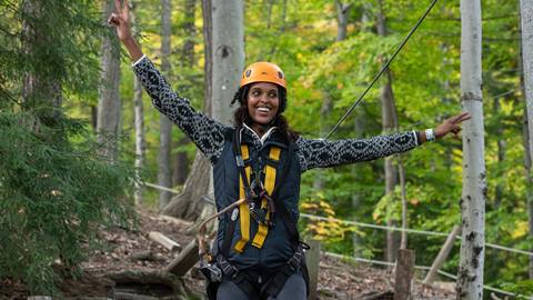 A woman in a helmet and harness smiles broadly while zip lining through a forest, arms outstretched and expressing joy.