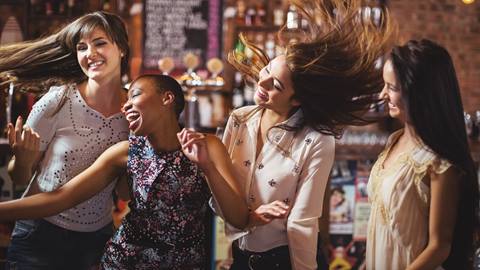 Four women laughing and dancing joyfully together in a bar with a warm, ambient atmosphere.