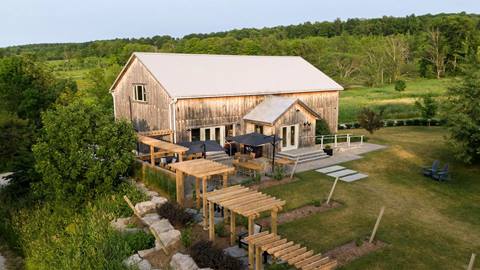 Aerial view of a rustic barn converted into a home with an adjoining outdoor patio and pergola, surrounded by lush greenery in a countryside setting.