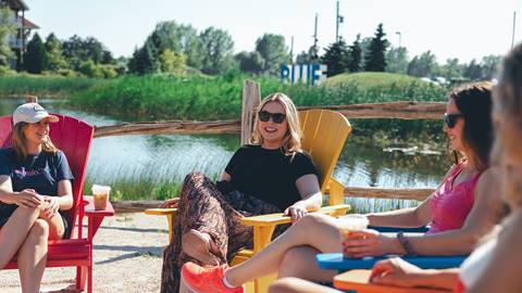 Three women relaxing in colorful chairs by a lake, chatting and enjoying drinks on a sunny day.
