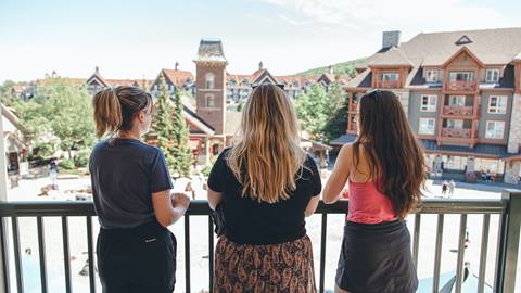 Three women standing on a balcony overlooking a bustling village square with buildings and trees in the background.