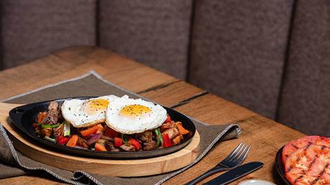 A skillet with sautéed vegetables and two fried eggs, served on a wooden table beside a grilled half tomato.