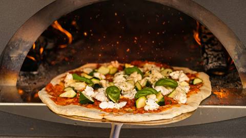 A pizza with basil and cheese toppings baking in a wood-fired oven, with visible flames on the side.