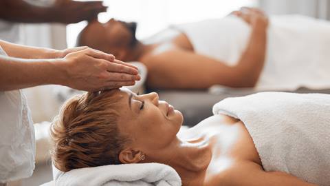 Two people receiving relaxing head massages in a serene spa environment, lying on treatment tables with therapists attending to them.