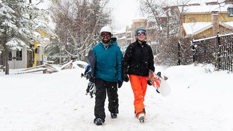 Two people walking down a snow covered street with snowboards.