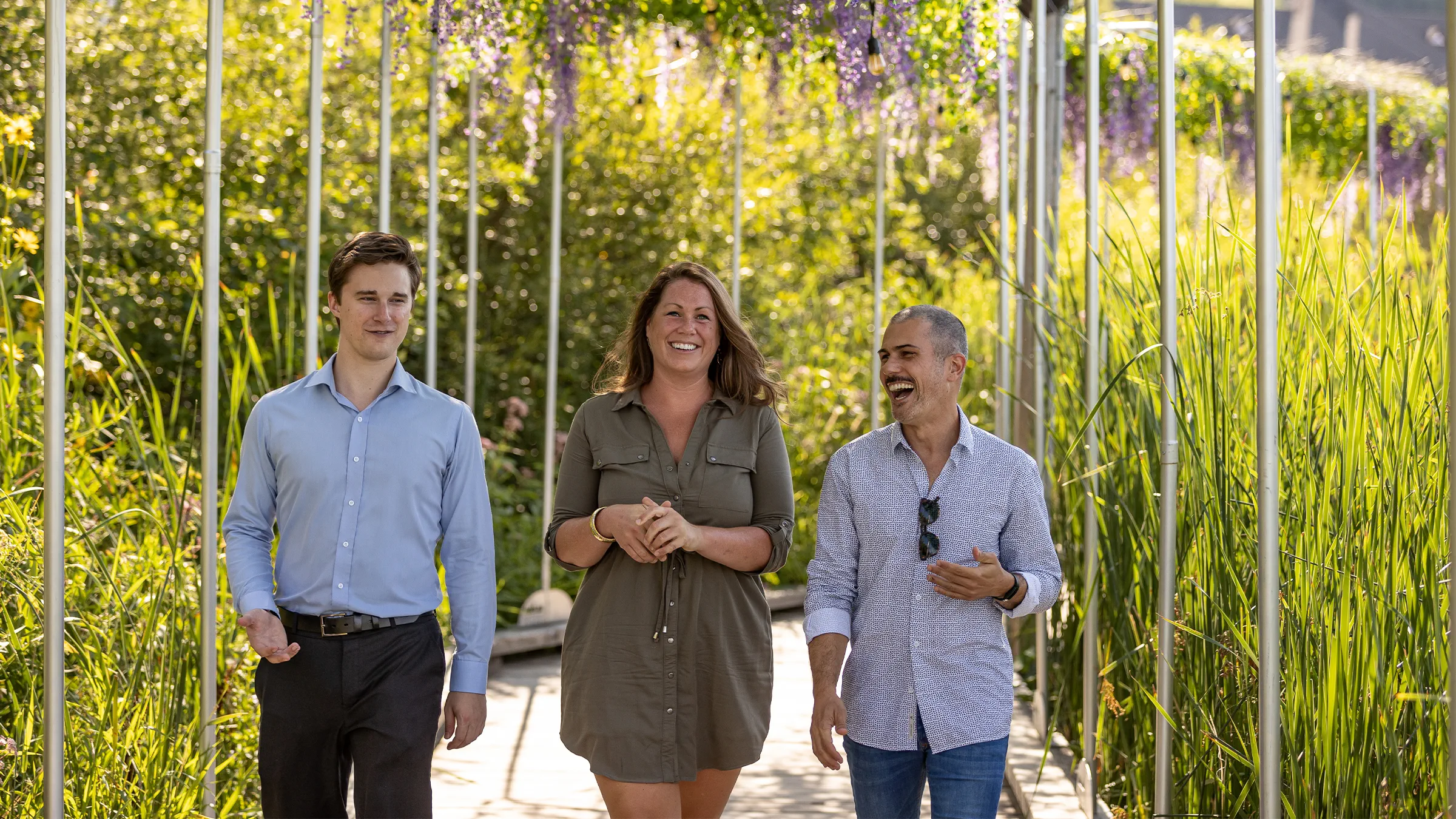 Three people walk together under a floral pergola in a garden.