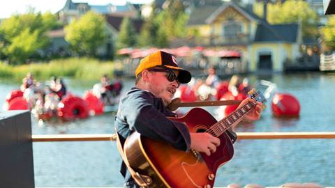 Musician playing guitar outdoors with pedal boats in the background.