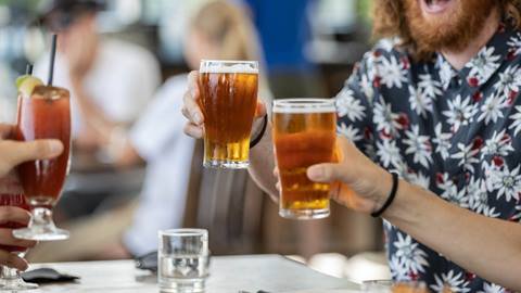 Three people clink their glasses—a Bloody Mary and two beers—at a casual gathering. Glasses of water are on the table.