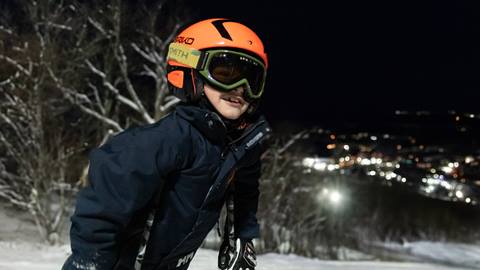 A young skier wearing an orange helmet and green goggles smiles on a snowy slope during night skiing, with blurred resort lights glowing in the background.