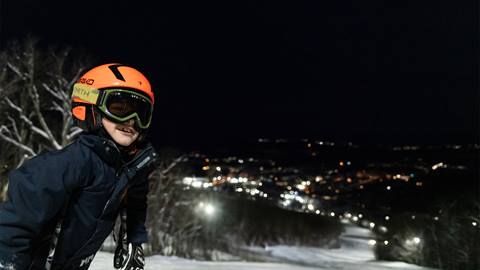 A young skier wearing an orange helmet and green goggles smiles on a snowy slope during night skiing, with blurred resort lights glowing in the background.