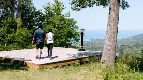 Two people walk hand in hand on a wooden platform surrounded by greenery, overlooking a scenic view of trees, water, and distant hills.