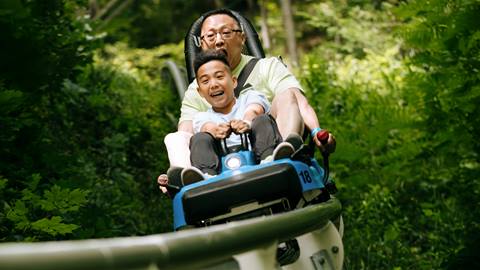 An adult and a child ride a blue roller coaster cart down a track surrounded by lush green foliage.