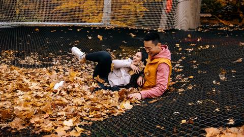 Two people sit and laugh together on a net covered with autumn leaves, surrounded by mesh fencing and yellow foliage.