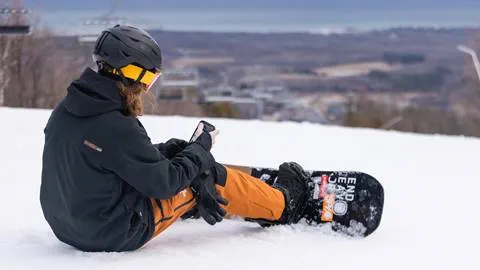 A snowboarder sitting on a snowy slope with a cell phone.