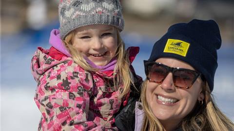A smiling child in a pink jacket held by an adult woman wearing sunglasses and a beanie outdoors.