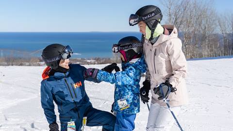 Three people in ski gear stand on a snowy hill overlooking a distant body of water and clear sky. They are wearing helmets and ski goggles.