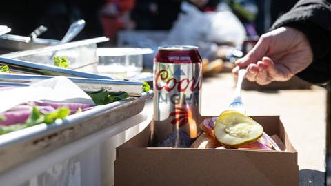 A person's hand picking up a pickle slice with a fork from a food container at an outdoor event with a canned beverage in the background.