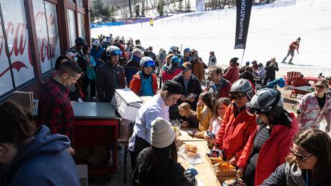 Skiers and snowboarders queuing for food at a busy outdoor resort eatery with a snowy mountainside in the background.