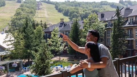 A couple stands on a balcony overlooking a resort with ski slopes and a pool, pointing at the scenery.