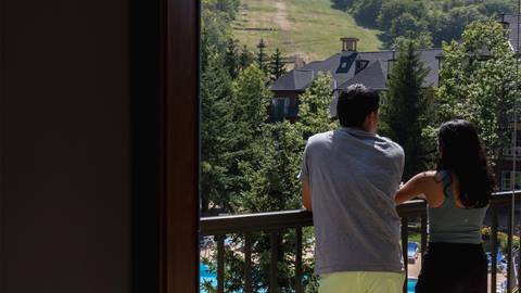 A man and woman stand on a balcony overlooking a wooded area with a pool below and a slope in the background on a sunny day.