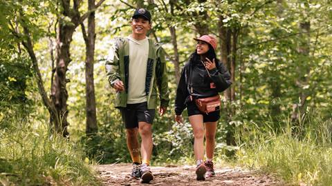 Two people walking on a forest trail. They are smiling and wearing casual outdoor clothing. Trees and greenery surround them.
