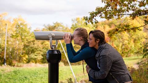 Two women looking through binoculars in a field.
