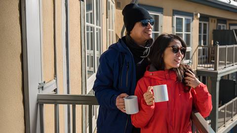 Two people wearing jackets and sunglasses hold mugs while standing on a balcony in the sunlight.