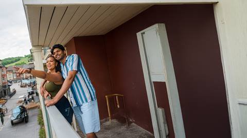 A smiling couple stands on a balcony overlooking a town square, with the woman pointing towards the buildings below.