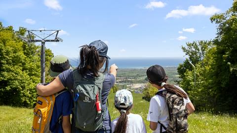 Four people with backpacks stand on a grassy hilltop looking out over a distant landscape, with one person pointing toward the horizon and a cable tower visible on the left.