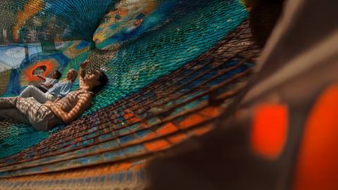 Three people relax on a colorful, netted structure, with dappled sunlight creating patterns on their bodies.