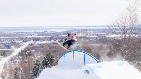 A snowboarder in mid-air performs a trick on a blue rail at a snowy terrain park with a town and water visible in the background.