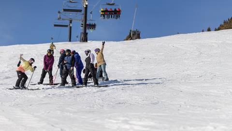 A group of skiers and snowboarders stand together on a snowy slope with a ski lift and blue sky in the background.