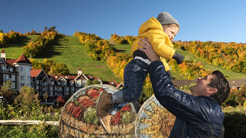 A smiling man lifting a toddler above a hay bale in an autumnal setting.