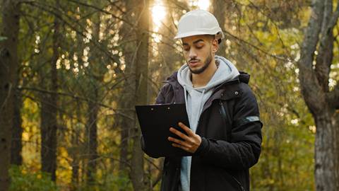 A man in a hard hat is holding a clipboard in the woods.