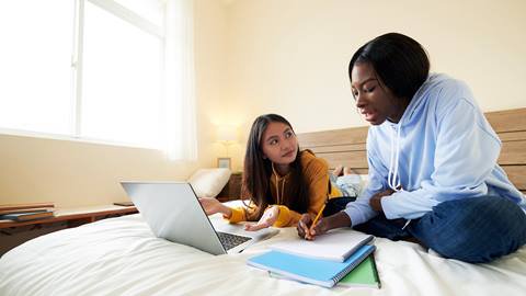 Two women working on a laptop on a bed.