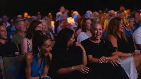 Audience members seated indoors, laughing and enjoying a performance, with dim lighting and others visible in the background.