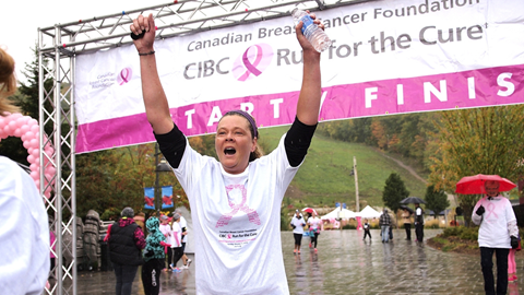 A person in a white shirt raises their arms in victory at the finish line of a "CIBC Run for the Cure" breast cancer awareness event.