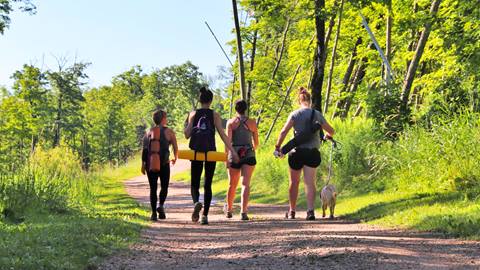 Four people walk along a dirt path in a forested area, with two carrying yoga mats and one walking a small dog on a leash.