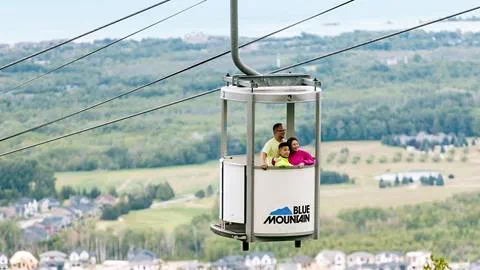 A cable car carrying two passengers ascends with the blue mountain logo visible, overlooking lush green landscapes below.