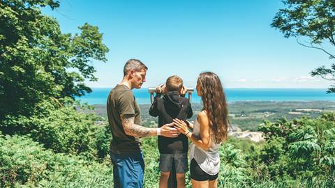 A family of three using binoculars to admire a panoramic view of a coastline from a forested overlook.